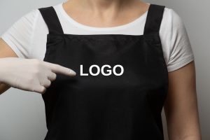 Closeup, fragment. Girl in black apron pointing finger to empty space on her chest. Protecting gloves. Safety concept. Copy space, place for text, mockup.